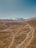 A vast and arid landscape extends towards rugged mountains capped with snow under a clear blue sky. The terrain is predominantly brown and barren, with winding dirt roads traversing through it. A small settlement can be seen in the middle of the plain, adding a hint of human presence in this natural setting.