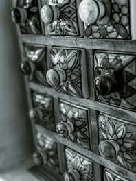 A chest of small, ornate drawers with ceramic knobs and intricately patterned surfaces. The drawers are arranged in a wooden frame, each with a unique floral or geometric design, contributing to an overall vintage and artistic appearance.