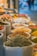 A vibrant market scene with large, open sacks filled with colorful spices and herbs. The sacks are stacked closely together, showcasing a variety of textures and colors. The blurred background suggests an indoor market environment, with a hint of other shops and products.