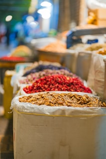 A variety of colorful dried items are displayed in large bags at a bustling market, with a focus on dried citrus slices and red spices. The background is softly blurred, giving a hint of more market stalls and people.