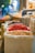 A variety of colorful dried items are displayed in large bags at a bustling market, with a focus on dried citrus slices and red spices. The background is softly blurred, giving a hint of more market stalls and people.
