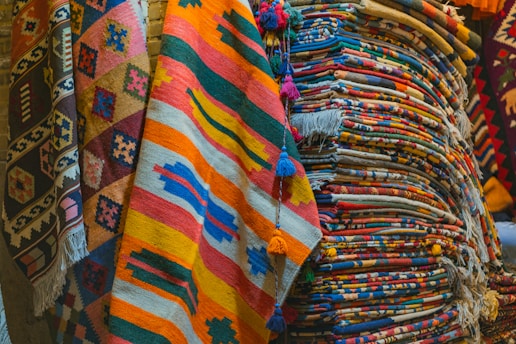 A collection of colorful woven rugs stacked and displayed. The rugs feature various intricate patterns and vibrant colors, including stripes, geometric shapes, and fringes. The textures appear rich and diverse, indicative of traditional craftsmanship.
