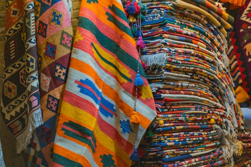 A collection of colorful woven rugs stacked and displayed. The rugs feature various intricate patterns and vibrant colors, including stripes, geometric shapes, and fringes. The textures appear rich and diverse, indicative of traditional craftsmanship.