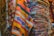 A collection of colorful woven rugs stacked and displayed. The rugs feature various intricate patterns and vibrant colors, including stripes, geometric shapes, and fringes. The textures appear rich and diverse, indicative of traditional craftsmanship.
