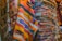 A collection of colorful woven rugs stacked and displayed. The rugs feature various intricate patterns and vibrant colors, including stripes, geometric shapes, and fringes. The textures appear rich and diverse, indicative of traditional craftsmanship.