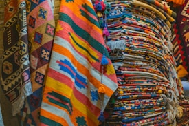 A collection of colorful woven rugs stacked and displayed. The rugs feature various intricate patterns and vibrant colors, including stripes, geometric shapes, and fringes. The textures appear rich and diverse, indicative of traditional craftsmanship.