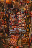 A vibrant display features an assortment of handmade textiles and bags adorned with colorful tassels. The scene is filled with rich patterns and textures, creating an intricate tapestry of colors. Pillows and bags are densely packed, each featuring a unique combination of geometric designs and bold hues.