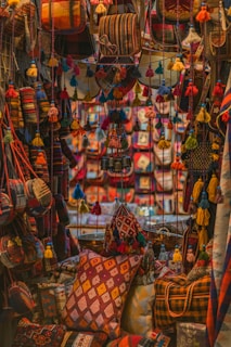 A vibrant display features an assortment of handmade textiles and bags adorned with colorful tassels. The scene is filled with rich patterns and textures, creating an intricate tapestry of colors. Pillows and bags are densely packed, each featuring a unique combination of geometric designs and bold hues.