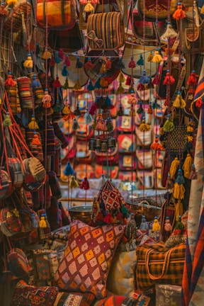 A vibrant display features an assortment of handmade textiles and bags adorned with colorful tassels. The scene is filled with rich patterns and textures, creating an intricate tapestry of colors. Pillows and bags are densely packed, each featuring a unique combination of geometric designs and bold hues.
