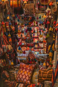 A vibrant display features an assortment of handmade textiles and bags adorned with colorful tassels. The scene is filled with rich patterns and textures, creating an intricate tapestry of colors. Pillows and bags are densely packed, each featuring a unique combination of geometric designs and bold hues.