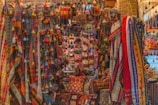 A vibrant display of colorful textiles, bags, and decorative items hanging and arranged on a market stall. The scene is filled with a variety of patterns and textures, featuring stripes, checks, and intricate designs in bright hues. Tassels and pom-poms add to the ornate look, creating a visually rich and lively atmosphere.