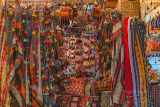 A vibrant display of colorful textiles, bags, and decorative items hanging and arranged on a market stall. The scene is filled with a variety of patterns and textures, featuring stripes, checks, and intricate designs in bright hues. Tassels and pom-poms add to the ornate look, creating a visually rich and lively atmosphere.