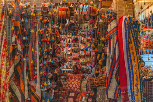 A vibrant display of colorful textiles, bags, and decorative items hanging and arranged on a market stall. The scene is filled with a variety of patterns and textures, featuring stripes, checks, and intricate designs in bright hues. Tassels and pom-poms add to the ornate look, creating a visually rich and lively atmosphere.