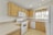 A bright kitchen with light wood cabinets and beige countertops. The kitchen includes a white gas stove, a microwave, and a dishwasher. There is a window above the sink with a colorful floral curtain. The room is well-lit with multiple recessed lights.