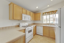 A bright kitchen with light wood cabinets and beige countertops. The kitchen includes a white gas stove, a microwave, and a dishwasher. There is a window above the sink with a colorful floral curtain. The room is well-lit with multiple recessed lights.