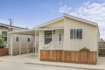 A small, single-story house with a beige exterior and a white door. There is a covered carport attached to the side and a small wooden fence in the front. The house has a simple, neat appearance with a few windows and some greenery in front.