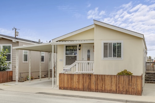 A small, single-story house with a beige exterior and a white door. There is a covered carport attached to the side and a small wooden fence in the front. The house has a simple, neat appearance with a few windows and some greenery in front.