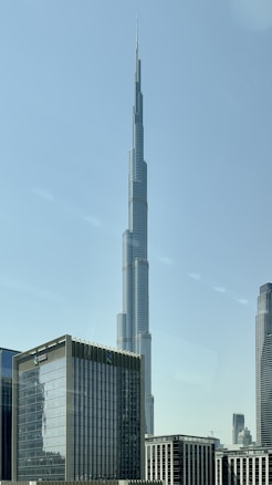 A tall skyscraper dominates the skyline, its sleek, futuristic design reaching into the clear blue sky. Surrounding the main tower are other modern office buildings, all with reflective glass facades, in an urban setting.