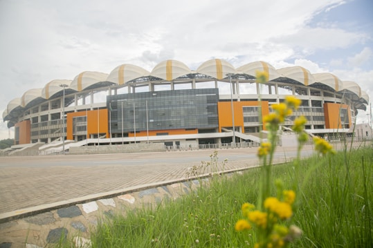 A large, modern stadium with an elaborate white and orange facade, featuring a distinct curved roof design. The structure dominates the background, while in the foreground, there are green grass and yellow flowers in focus, adding a natural element to the scene.