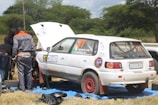 A group of people is working on a white rally car with red rims parked on a blue tarp in a grassy area. One person is wearing a gray and orange jumpsuit with the text 'AGRO AUTO SPAR' on the back. The car's hood is open, and it has several stickers and numbers, including '27' and 'ZAMBIA Rally Championship'. Trees and a cloudy sky are in the background.