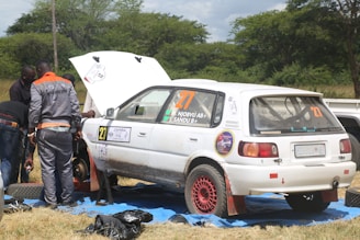 A group of people is working on a white rally car with red rims parked on a blue tarp in a grassy area. One person is wearing a gray and orange jumpsuit with the text 'AGRO AUTO SPAR' on the back. The car's hood is open, and it has several stickers and numbers, including '27' and 'ZAMBIA Rally Championship'. Trees and a cloudy sky are in the background.