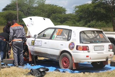 A group of people is working on a white rally car with red rims parked on a blue tarp in a grassy area. One person is wearing a gray and orange jumpsuit with the text 'AGRO AUTO SPAR' on the back. The car's hood is open, and it has several stickers and numbers, including '27' and 'ZAMBIA Rally Championship'. Trees and a cloudy sky are in the background.