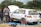 A group of people is working on a white rally car with red rims parked on a blue tarp in a grassy area. One person is wearing a gray and orange jumpsuit with the text 'AGRO AUTO SPAR' on the back. The car's hood is open, and it has several stickers and numbers, including '27' and 'ZAMBIA Rally Championship'. Trees and a cloudy sky are in the background.