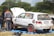 A group of people is working on a white rally car with red rims parked on a blue tarp in a grassy area. One person is wearing a gray and orange jumpsuit with the text 'AGRO AUTO SPAR' on the back. The car's hood is open, and it has several stickers and numbers, including '27' and 'ZAMBIA Rally Championship'. Trees and a cloudy sky are in the background.