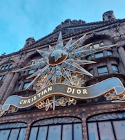 A grand architectural facade features an elaborate decorative display with a prominent star and floral design. It incorporates a large globe emblem at the center. Below the star, a banner with the words 'Christian Dior' is elegantly draped. The building is adorned with numerous lights, creating a striking and glamorous appearance against the blue sky.