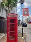 A red telephone booth labeled 'Taiping' stands on a sidewalk next to a lamppost with a sign that says 'Selamat Datang'. In the background, there are modern buildings, and a few cars are parked along the street. A tall palm tree is near the booth, adding a tropical element to the urban setting.