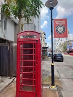 A red telephone booth labeled 'Taiping' stands on a sidewalk next to a lamppost with a sign that says 'Selamat Datang'. In the background, there are modern buildings, and a few cars are parked along the street. A tall palm tree is near the booth, adding a tropical element to the urban setting.