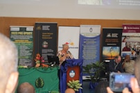 A conference or seminar setting with a speaker at a podium adorned with a blue cloth and floral arrangement. Behind the speaker are several banners and posters featuring organizations such as the Australian National University and themes like development policy. Attendees are seated in the foreground, one taking a photo with a phone.