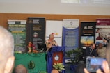 A conference or seminar setting with a speaker at a podium adorned with a blue cloth and floral arrangement. Behind the speaker are several banners and posters featuring organizations such as the Australian National University and themes like development policy. Attendees are seated in the foreground, one taking a photo with a phone.