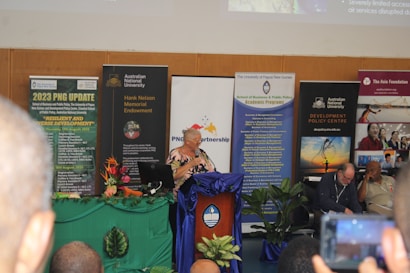 A conference or seminar setting with a speaker at a podium adorned with a blue cloth and floral arrangement. Behind the speaker are several banners and posters featuring organizations such as the Australian National University and themes like development policy. Attendees are seated in the foreground, one taking a photo with a phone.