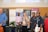 A group of people is gathered in an indoor setting, standing in front of several promotional banners. They appear to be posing for a photo, and the atmosphere seems to be casual and relaxed. One person is holding a notebook, and there are plants in the foreground.