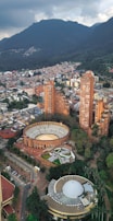 Aerial view of a cityscape featuring a prominent circular bullring and several tall residential buildings surrounded by lush greenery. In the background, a large mountain range looms under a cloudy sky. The architecture includes a mix of modern and historic styles.