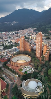 Aerial view of a cityscape featuring a prominent circular bullring and several tall residential buildings surrounded by lush greenery. In the background, a large mountain range looms under a cloudy sky. The architecture includes a mix of modern and historic styles.