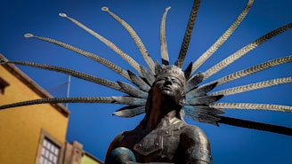 A bronze statue of a figure wearing an ornate headdress with multiple long feathers, set against a clear blue sky. The figure's expression is contemplative, and the background includes part of a building with a yellow facade.