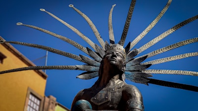 A bronze statue of a figure wearing an ornate headdress with multiple long feathers, set against a clear blue sky. The figure's expression is contemplative, and the background includes part of a building with a yellow facade.