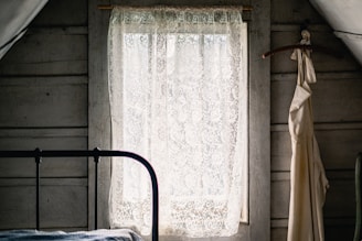 Sunlit vintage farmhouse bedroom with lace curtains and distressed wooden furniture.