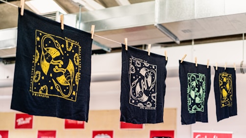 A series of intricately designed fabric prints hang on a line, each featuring unique designs in gold, white, and green against dark backgrounds. The prints are secured with wooden clothespins and appear to be in a workshop or studio environment with exposed ductwork visible above.