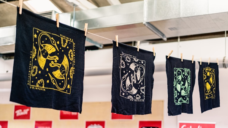 A series of intricately designed fabric prints hang on a line, each featuring unique designs in gold, white, and green against dark backgrounds. The prints are secured with wooden clothespins and appear to be in a workshop or studio environment with exposed ductwork visible above.