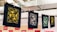 A series of intricately designed fabric prints hang on a line, each featuring unique designs in gold, white, and green against dark backgrounds. The prints are secured with wooden clothespins and appear to be in a workshop or studio environment with exposed ductwork visible above.
