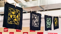 A series of intricately designed fabric prints hang on a line, each featuring unique designs in gold, white, and green against dark backgrounds. The prints are secured with wooden clothespins and appear to be in a workshop or studio environment with exposed ductwork visible above.
