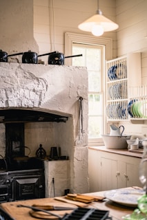 A rustic kitchen with a vintage, white-painted brick stove and various black pots sitting on top. The kitchen is adorned with open shelving displaying colorful plates, and a window lets in natural light. There's a pitcher and bowl set on the wooden countertop and utensils placed on the table in the foreground.