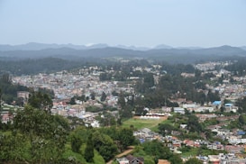 A panoramic view of a cityscape nestled among lush green hills and dense forests. The landscape features a patchwork of colorful buildings and residential areas, interconnected by winding roads. In the distance, rolling hills and mountains create a serene backdrop. The city appears lively with a blend of urban development and natural beauty.
