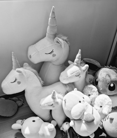 A collection of plush toys featuring several unicorns with closed eyes and heart-shaped details on their cheeks. Surrounding the unicorns are other plush toys, including a round bear and other animal figures. The toys are arranged closely together on a wooden floor.