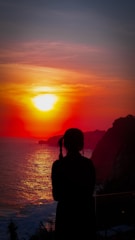 A silhouette of a person with braided hair stands by the sea, viewing a vibrant sunset. The sun casts a warm glow over the ocean, and rugged cliffs are visible in the background.