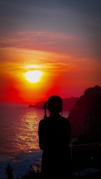 A silhouette of a person with braided hair stands by the sea, viewing a vibrant sunset. The sun casts a warm glow over the ocean, and rugged cliffs are visible in the background.
