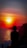 A silhouette of a person with braided hair stands by the sea, viewing a vibrant sunset. The sun casts a warm glow over the ocean, and rugged cliffs are visible in the background.